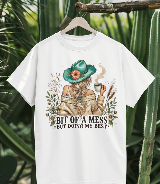 White t-shirt with a graphic design of a person in a hat and scarf, surrounded by plants, hanging against a cactus background.