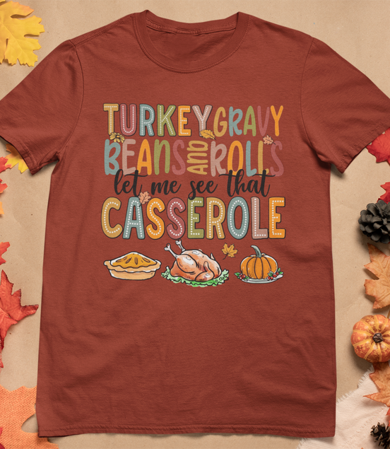 Brown t-shirt with colorful Thanksgiving-themed text and graphics on a beige background with fall decorations.