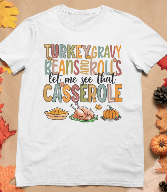 White t-shirt with Thanksgiving-themed text and graphics on a brown background with fall decorations.