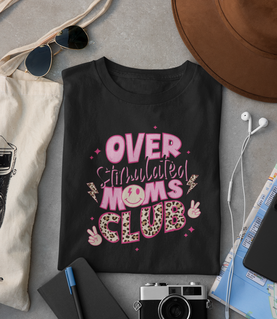 Black t-shirt with 'Overstimulated Moms Club' text, surrounded by sunglasses, camera, hat, and other items on a gray surface.