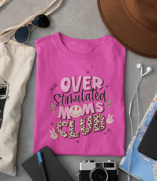 Pink t-shirt with 'Overstimulated Moms Club' text on a gray surface with accessories.