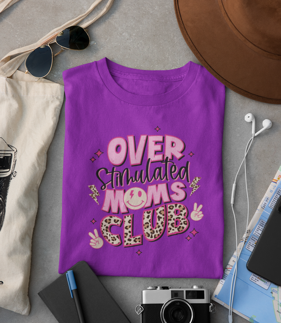 Purple t-shirt with 'Over Stimulated Moms Club' text on a gray surface with accessories.