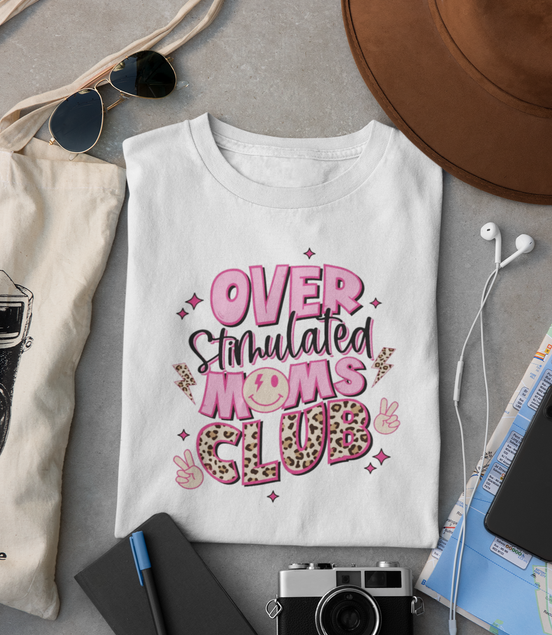 White t-shirt with 'Overstimulated Moms Club' text on a gray surface with accessories.