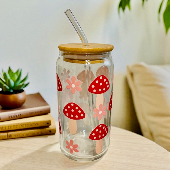 Clear tumbler with red mushroom and flower pattern, wooden lid, and straw on a table.