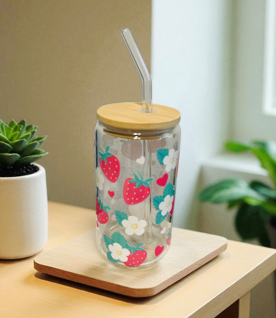Clear tumbler with strawberry and flower pattern on a wooden coaster, surrounded by plants.