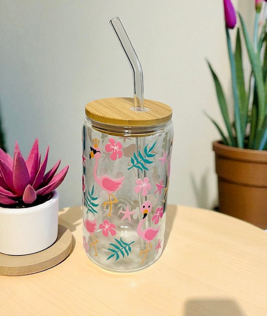 Glass tumbler with flamingo design, wooden lid, and straw on a table with plants in the background